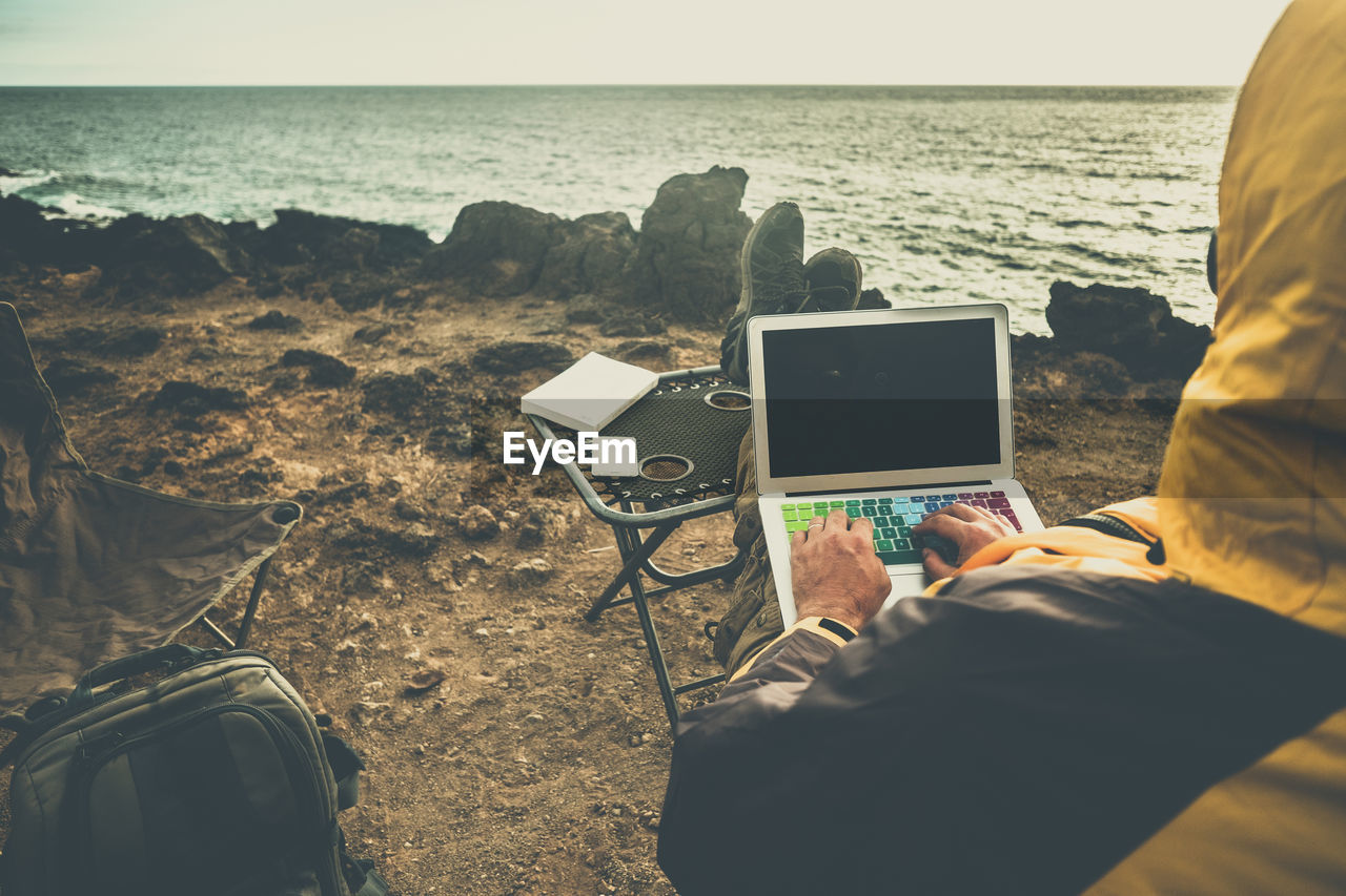 Rear view of man using laptop while sitting at beach against sea 