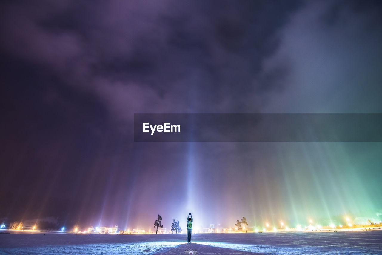 Rear view of person with arms raised standing at illuminated beach against sky at night