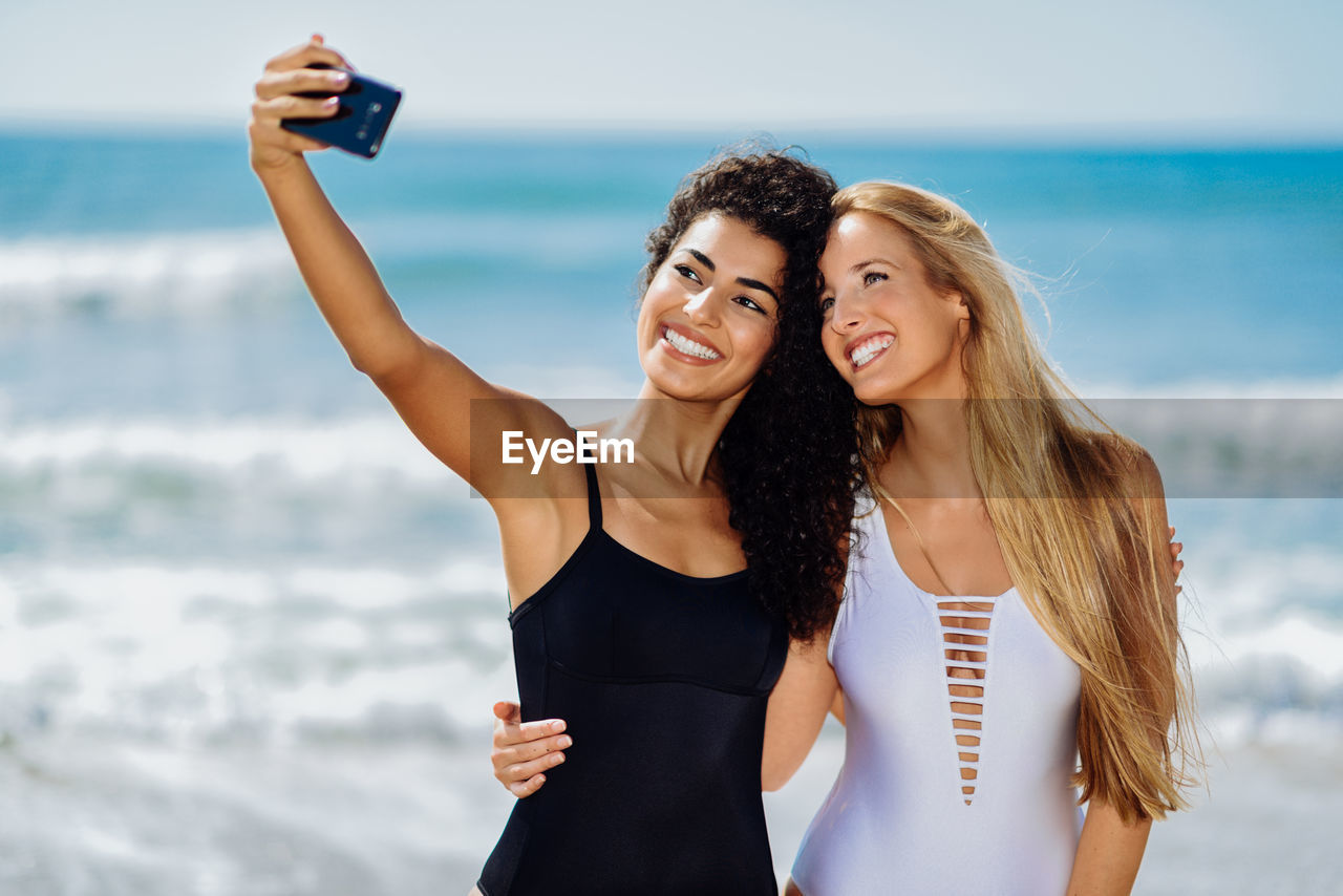 Happy female friends taking selfie while standing at beach