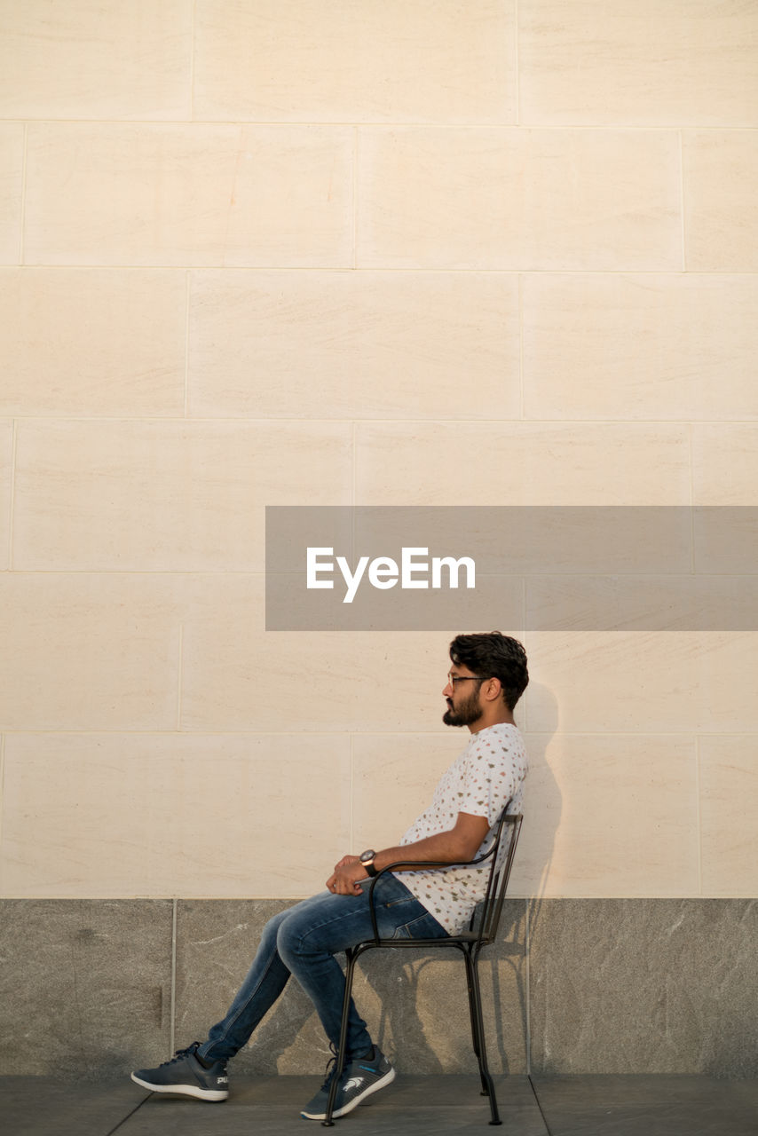 Side view of man sitting on chair against wall