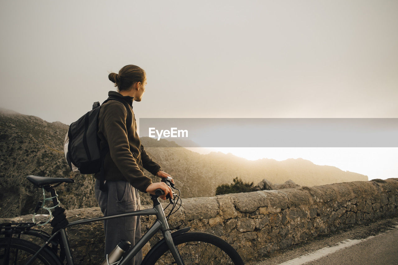 Side view of young man looking at mountains while standing with bicycle on road