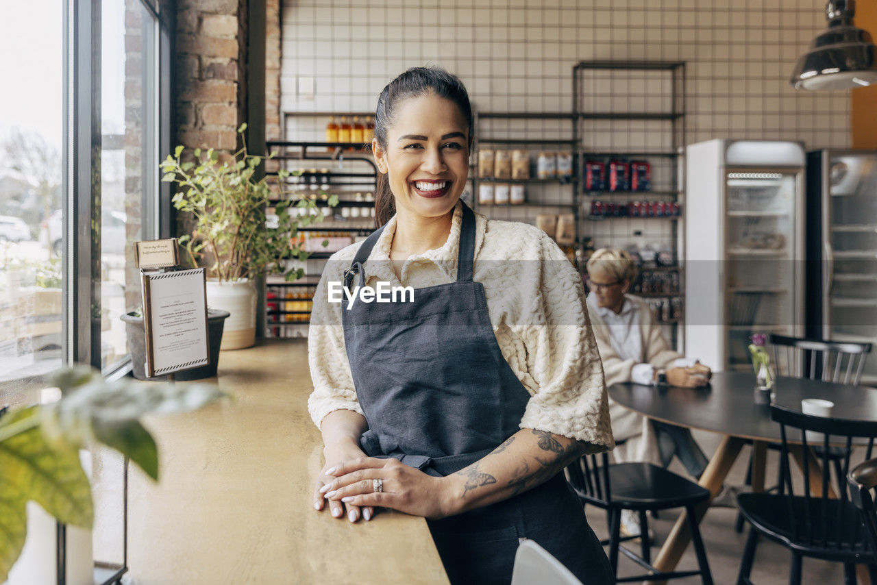 Portrait of happy female owner with tattooed arm standing near table in cafe