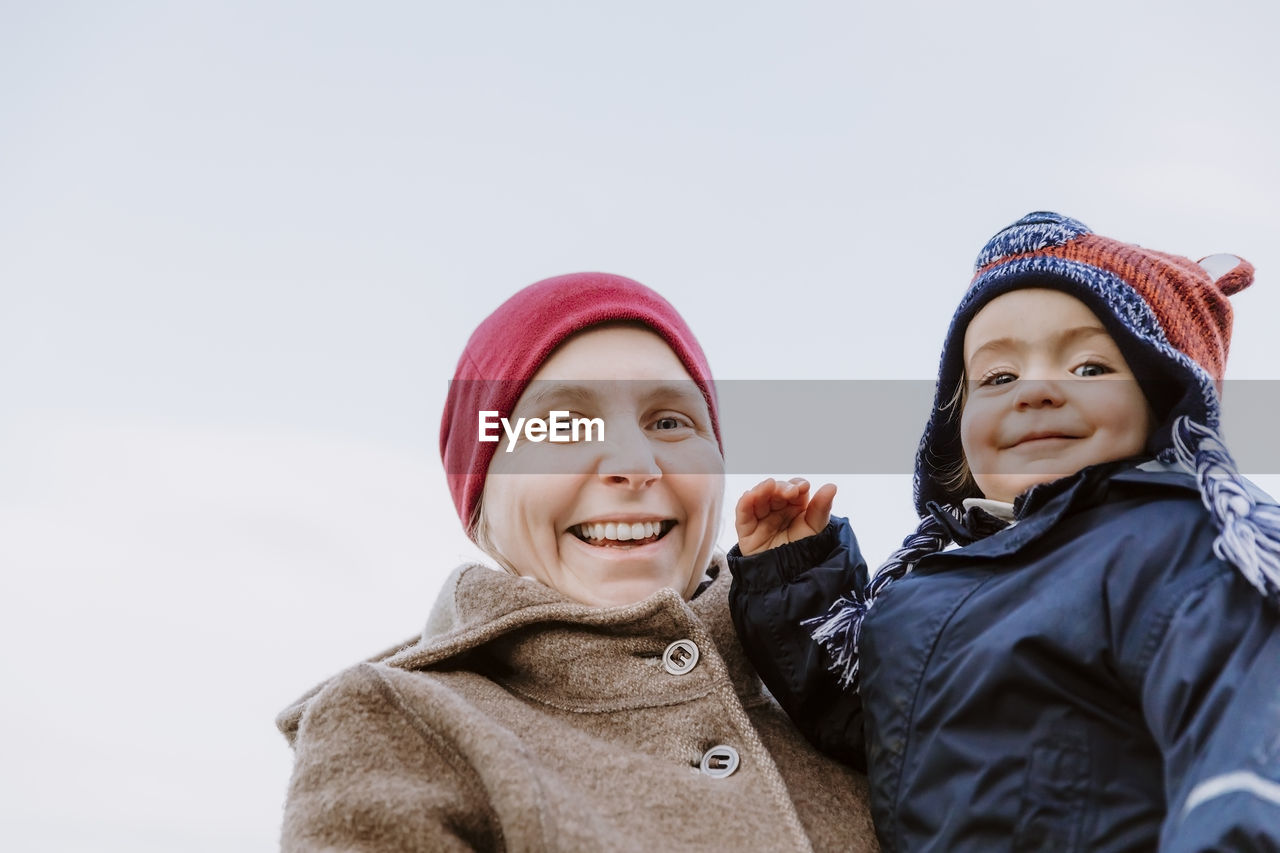 Portrait of smiling woman carrying daughter against sky during winter