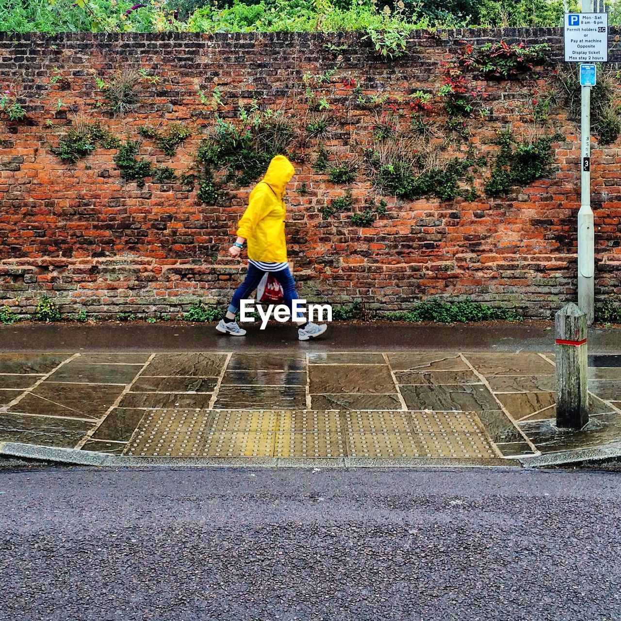 Woman wearing raincoat walking wet sidewalk by brick wall