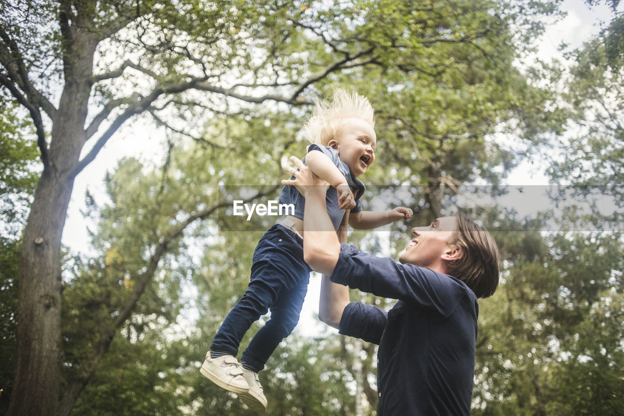 Low angle view of smiling man catching blond baby boy against trees