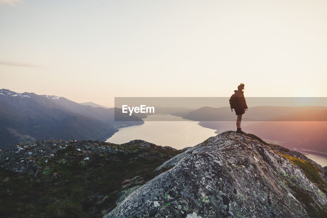 Rear view of woman looking at river amidst mountains against sky during sunset