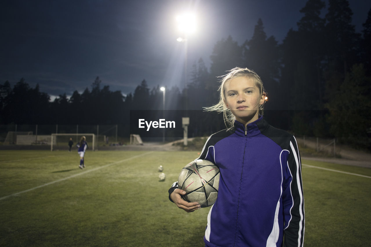 Portrait of girl standing with soccer ball on field against trees at night