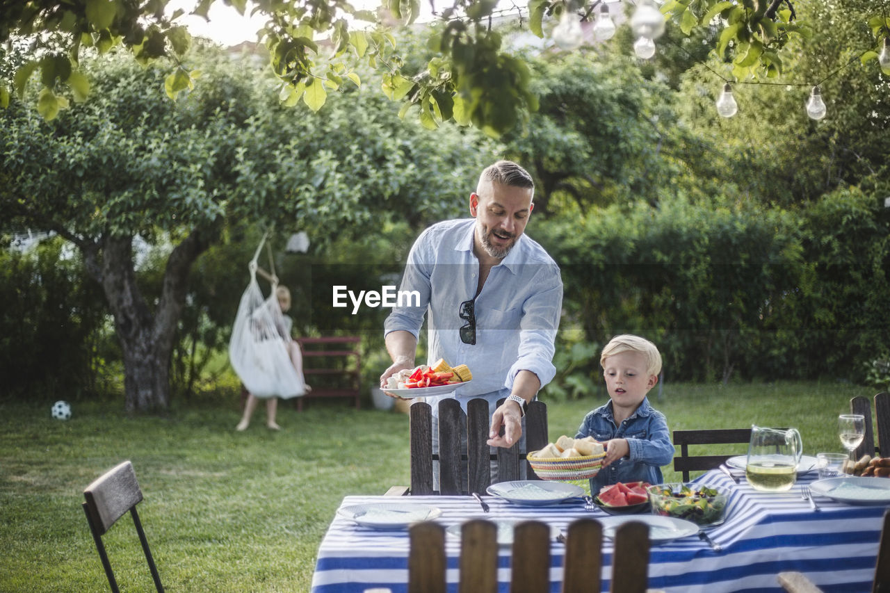 Mature father directing son with food plate while pointing at table in yard