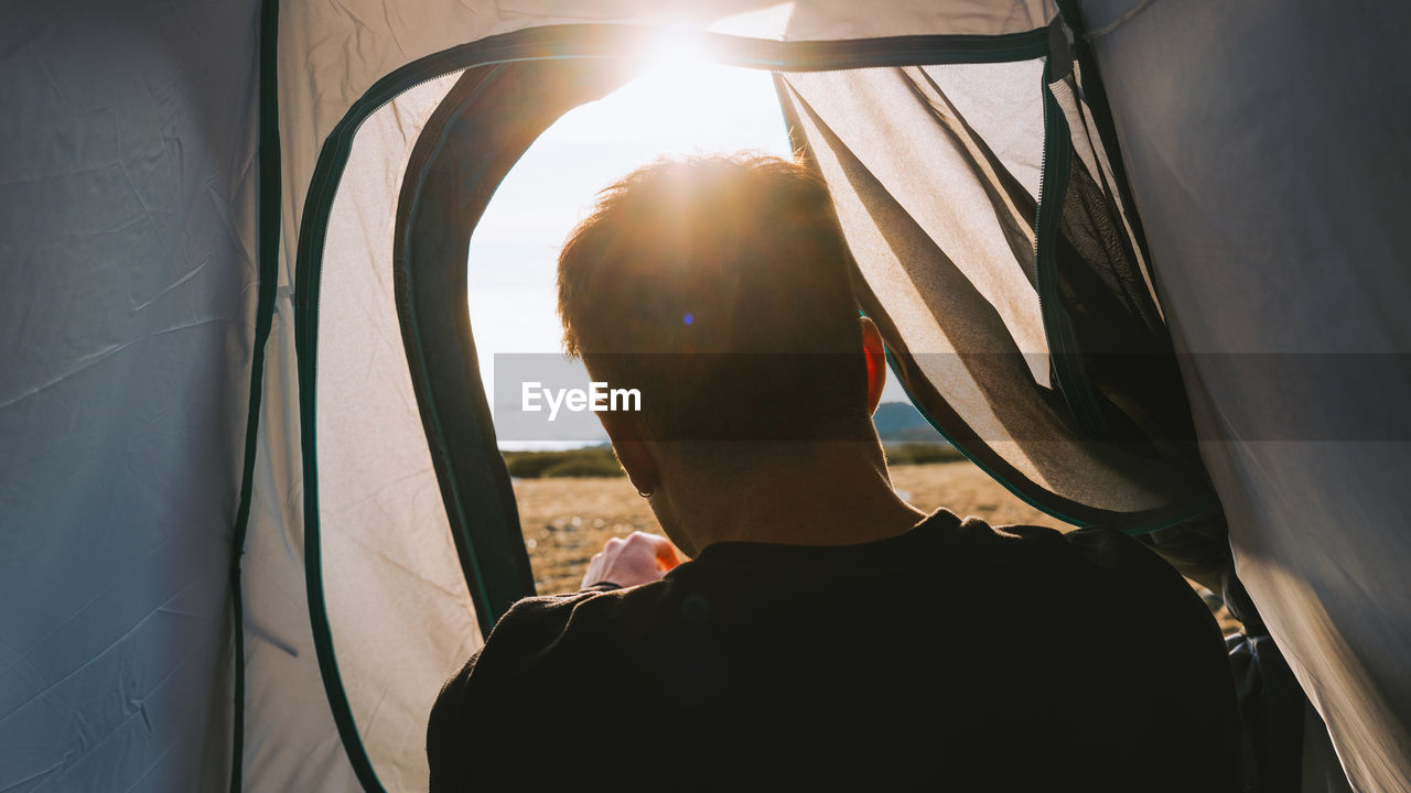 Back view of crop unrecognizable male traveler in casual clothes lying in camping tent and admiring picturesque mountain landscape on sunny day