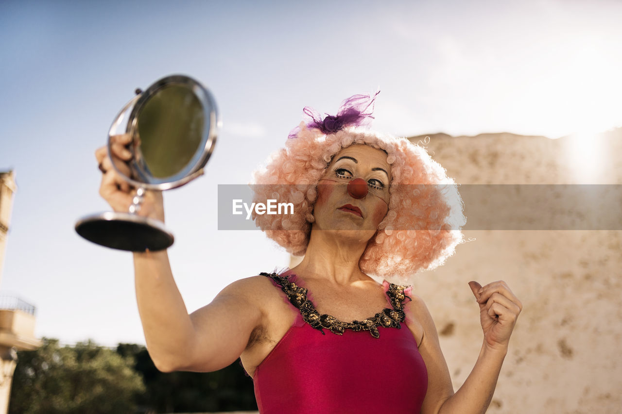 Confident mature female clown in bright costume and funny curly wig with makeup looking in round cosmetic mirror while standing on street on sunny day