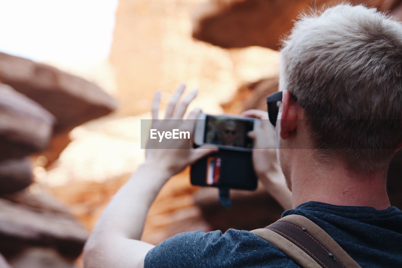 Rear view of man by rock formation taking selfie from mobile phone