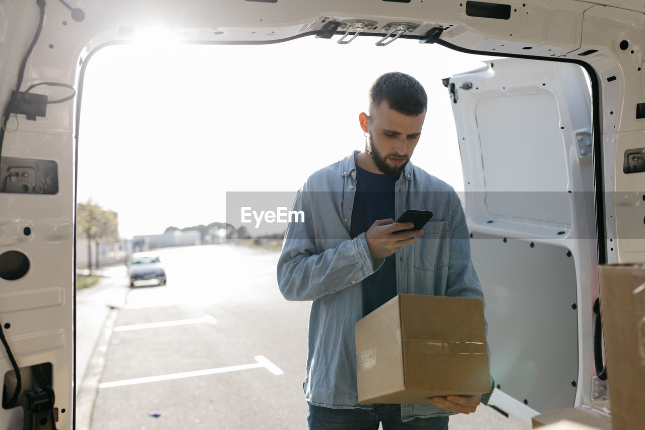 Delivery man using mobile phone holding box at van doorway