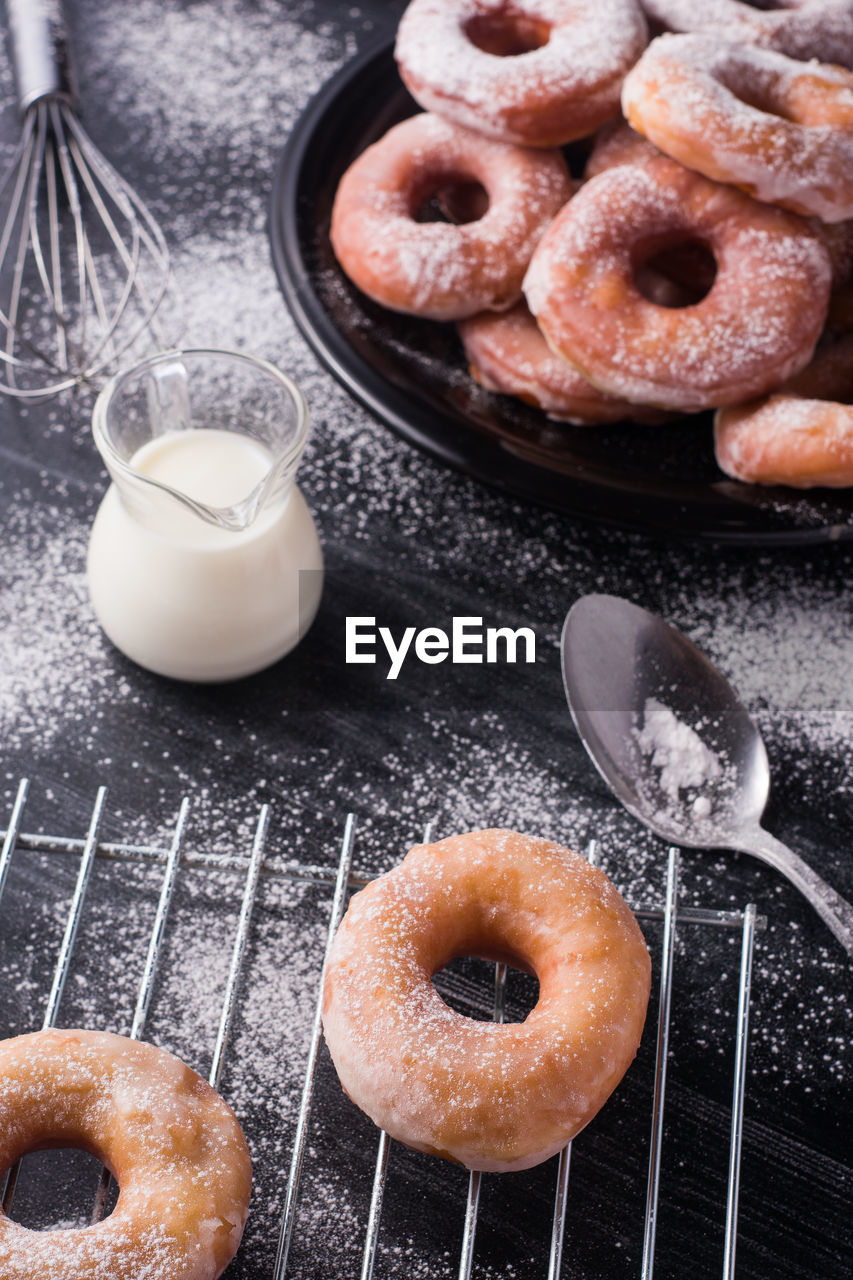 Sweet fried doughnuts served on plate near metal cooling rack and jug of milk on black messy table with powdered sugar