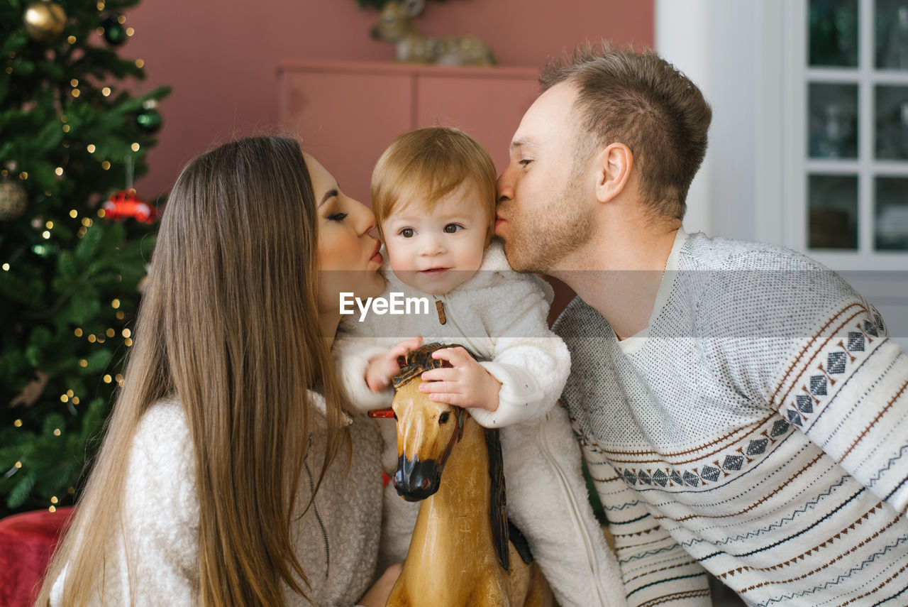 A small child of one year is sitting on a wooden horse near the christmas tree