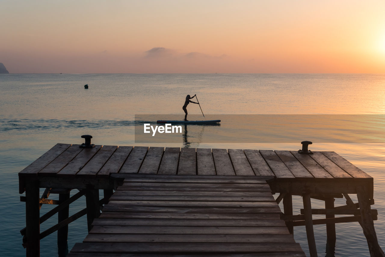 Side view of silhouette of unrecognizable female surfing on paddledboard on calm sea water at sunset in summer