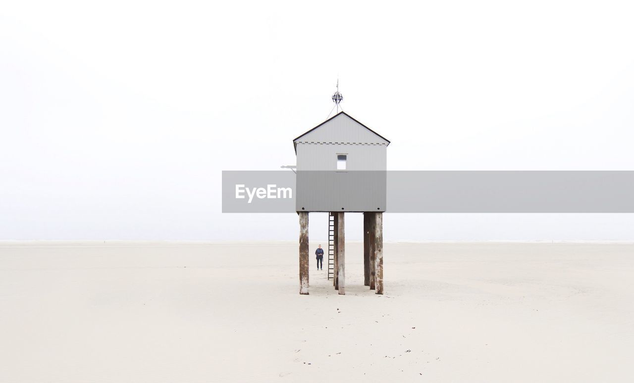 Woman standing by stilt cottage at beach against clear sky