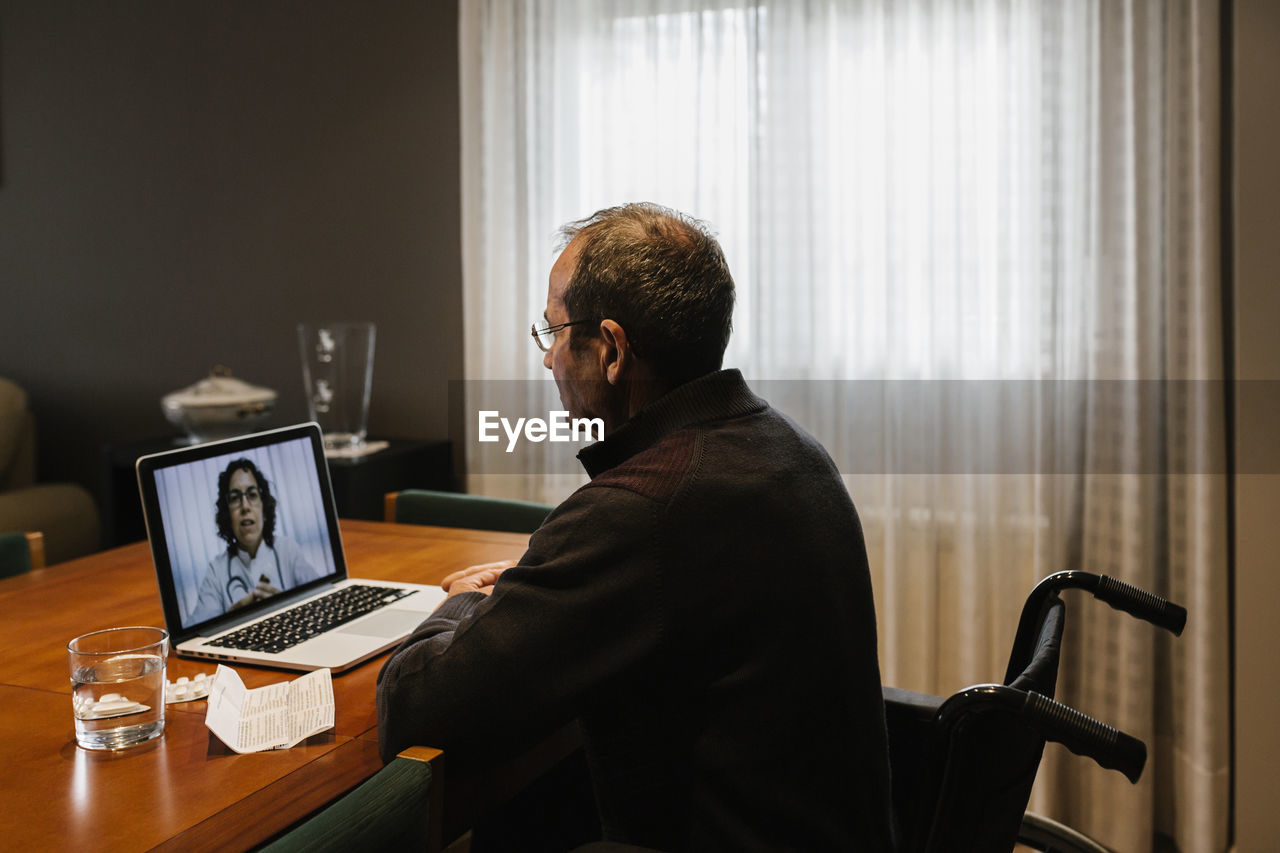 Disabled senior man sitting on wheelchair while female doctor consulting through video call on laptop