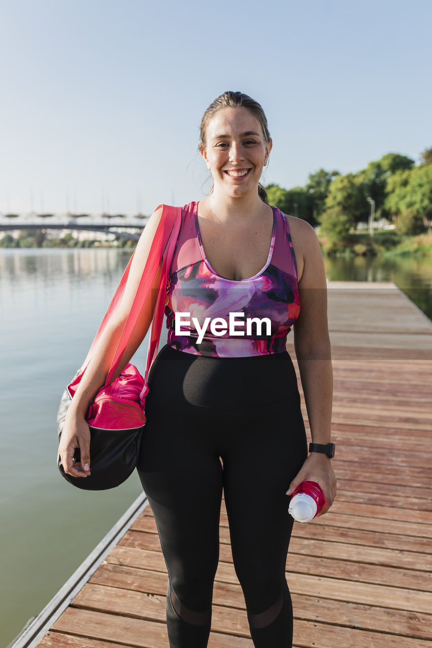 Smiling sportswoman with bag standing on pier