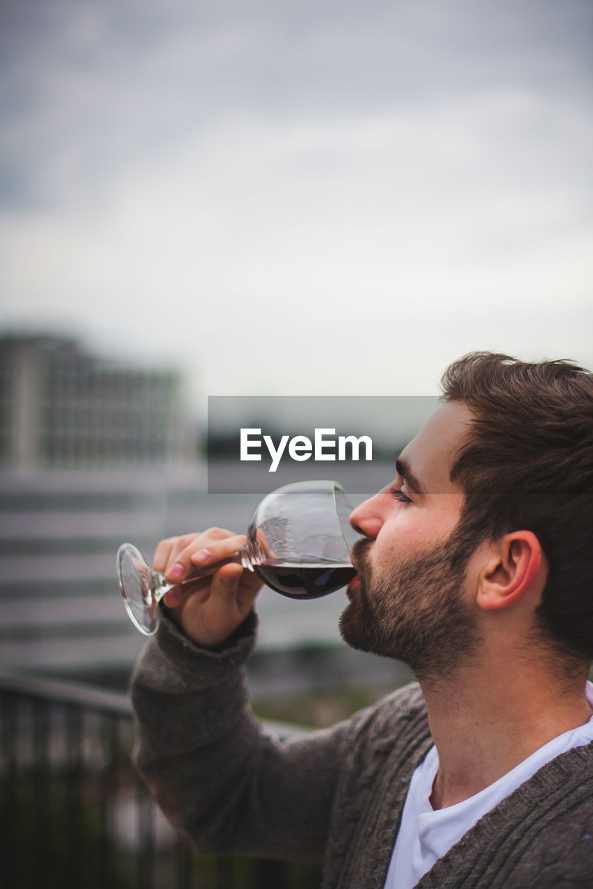 Side view of young man drinking red wine against sky
