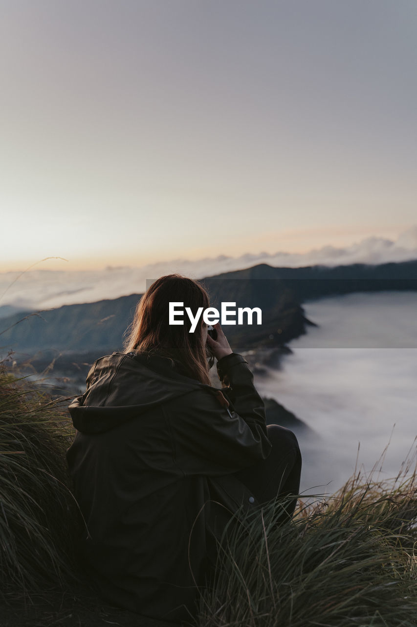 Side view of woman photographing sea while sitting on cliff against sky during sunset