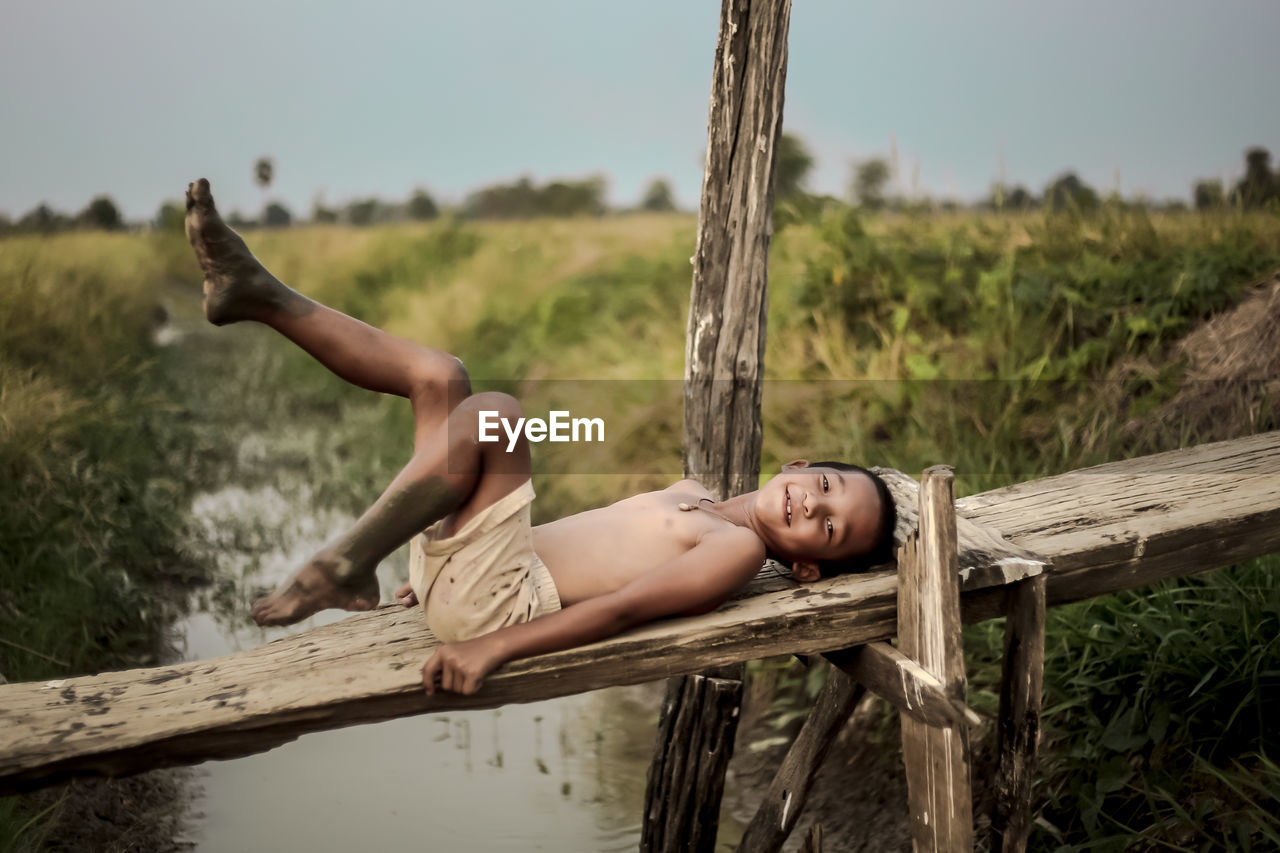 Portrait of shirtless boy lying on wood at farm