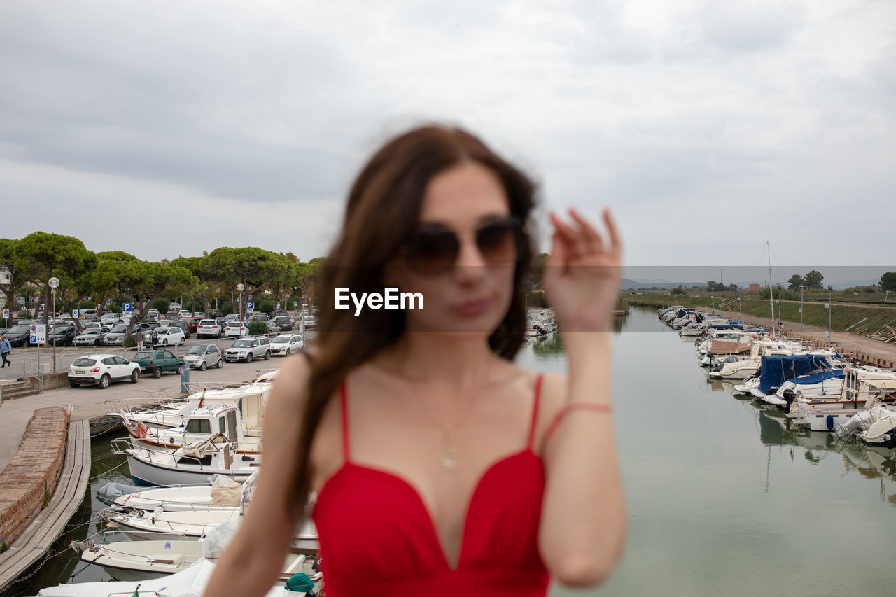 Portrait of young woman standing against nautical vessels moored by lake against sky