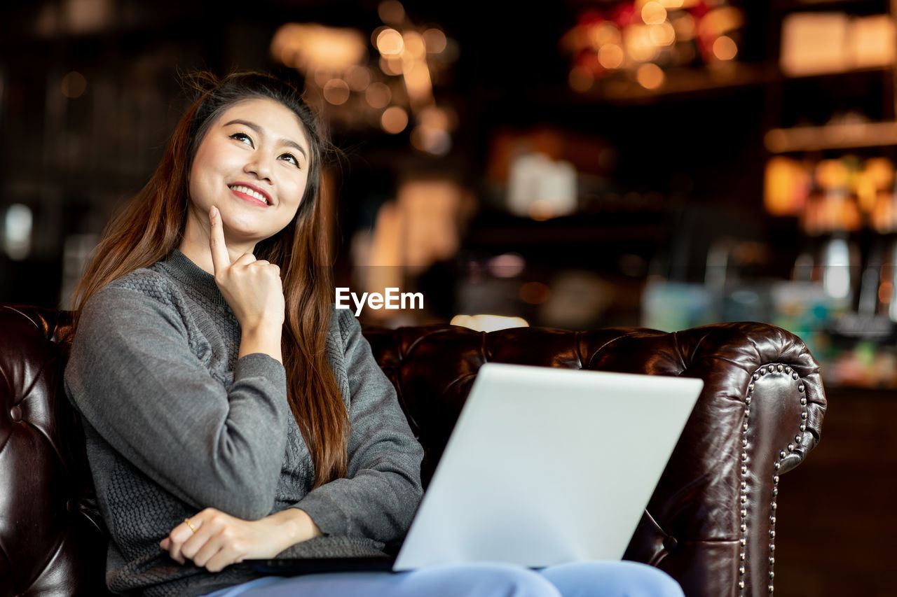 Thoughtful smiling young woman with laptop sitting on armchair