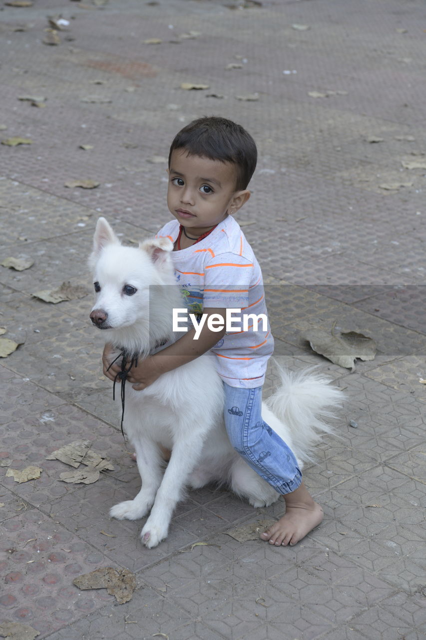 Portrait of cute baby boy sitting on a dog on the footpath and holding the dog with his neck