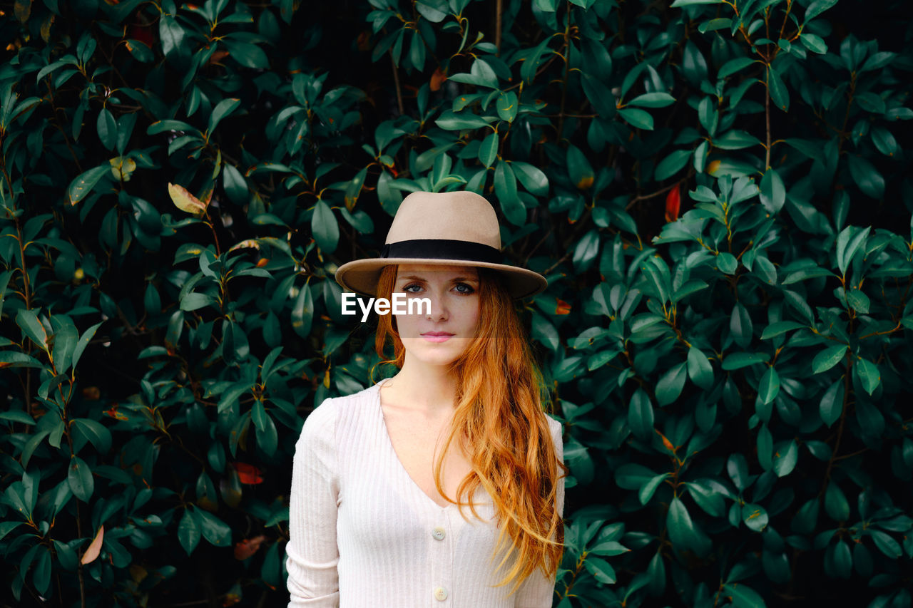 Attractive young long haired ginger female in stylish hat looking at camera while standing against green bushes in summer garden