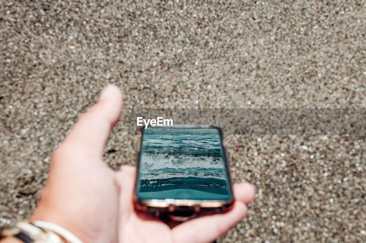 Close-up of hand holding smart phone reflected sea against beach