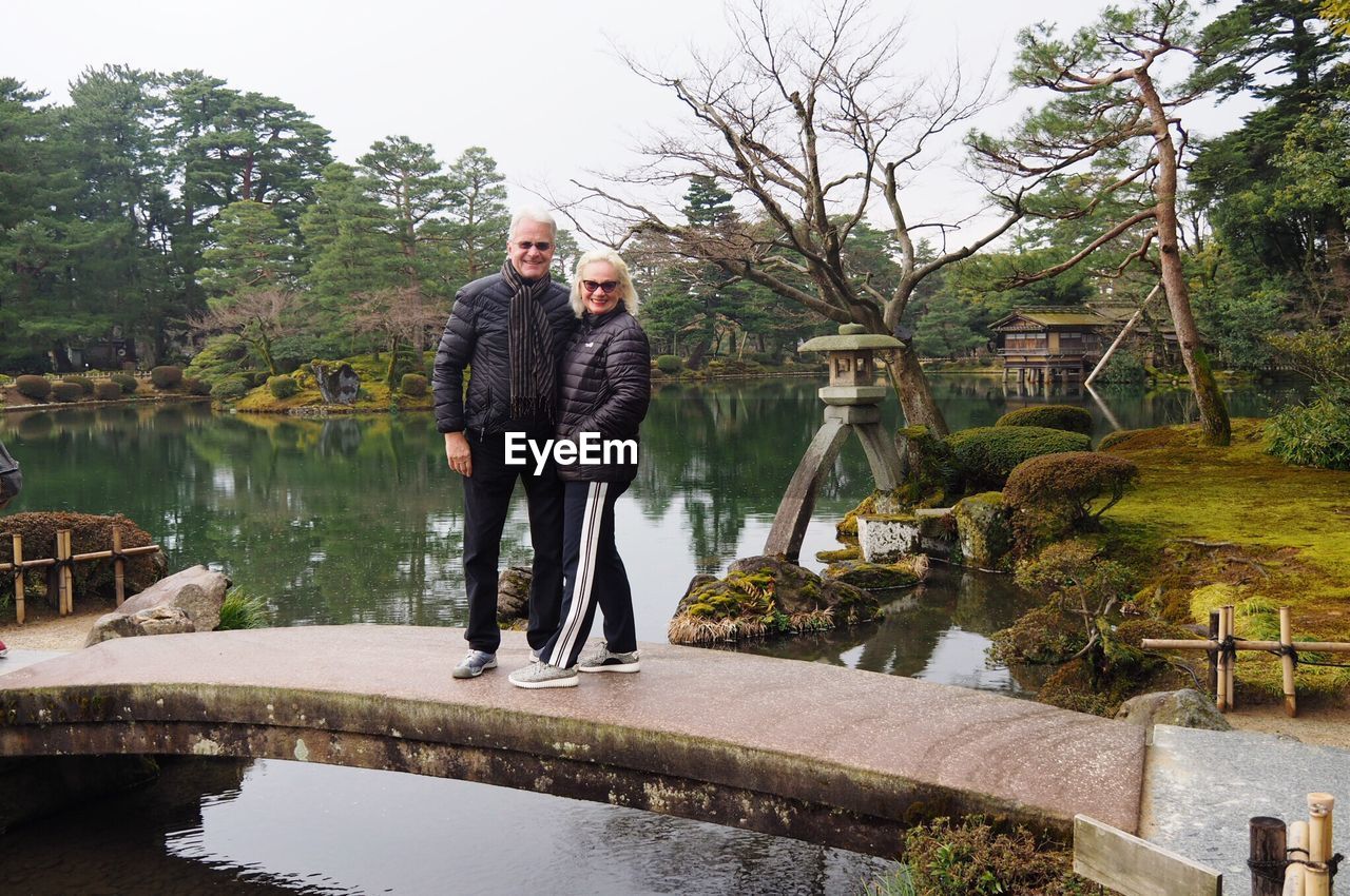 Portrait of happy mature couple standing on footbridge over pond at kenroku-en