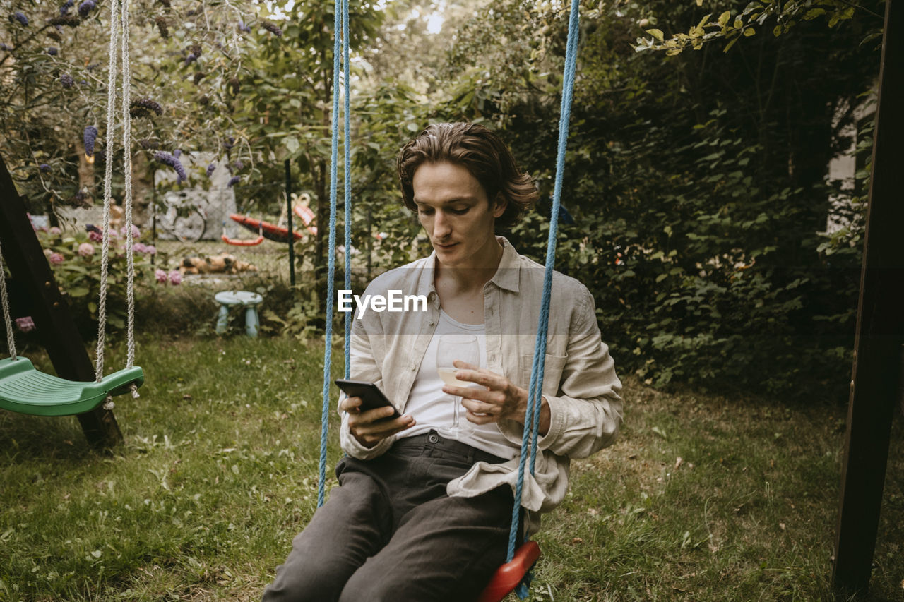Contemplating man with drink using smart phone while sitting on swing in yard