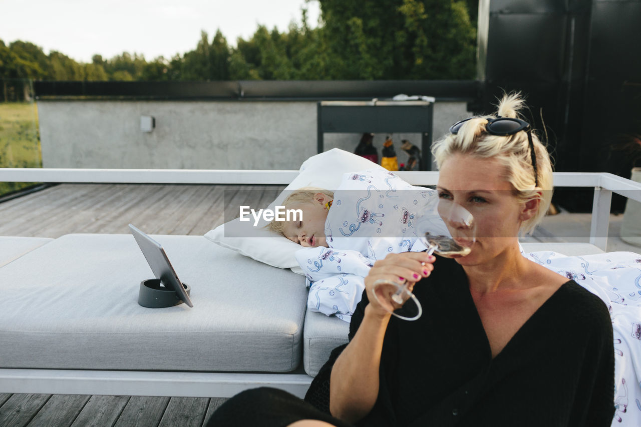 Mother relaxing with glass of wine while girl sleeping on terrace
