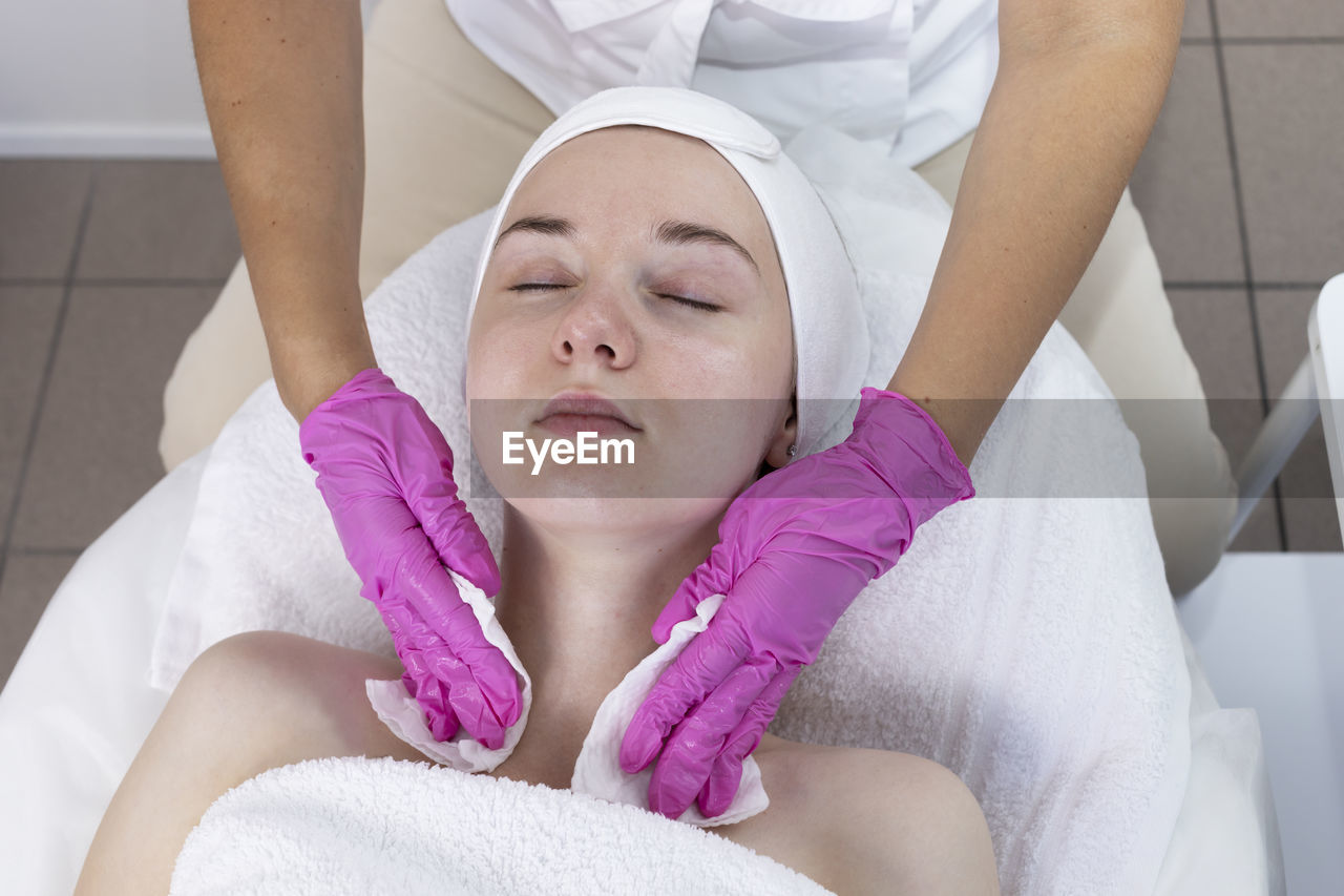 Top view cosmetologist clears skin of neck and face of young woman before beauty procedure