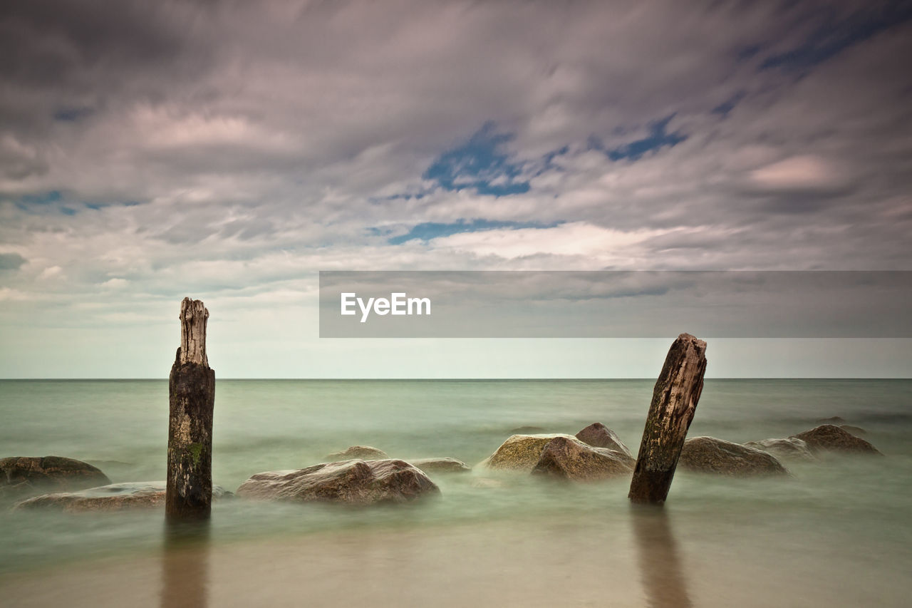 Weathered wooden posts by rocks in sea against cloudy sky