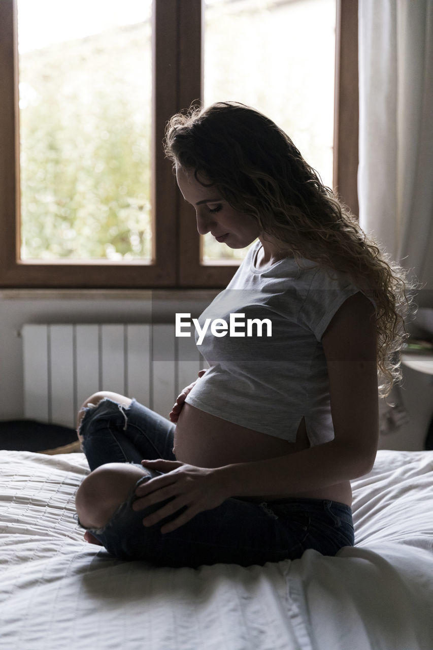Young pregnant woman looking at abdomen while sitting on bed