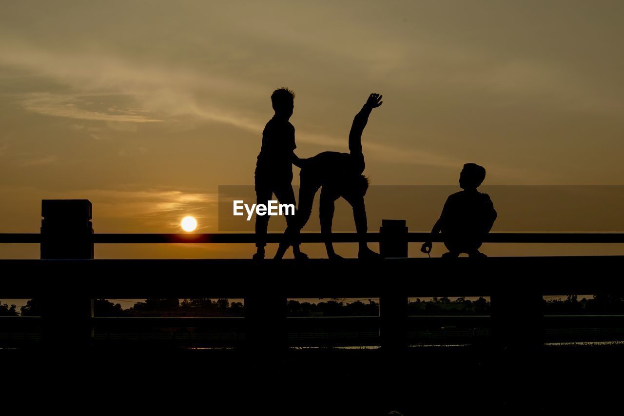 Silhouette male friends on railing against orange sky