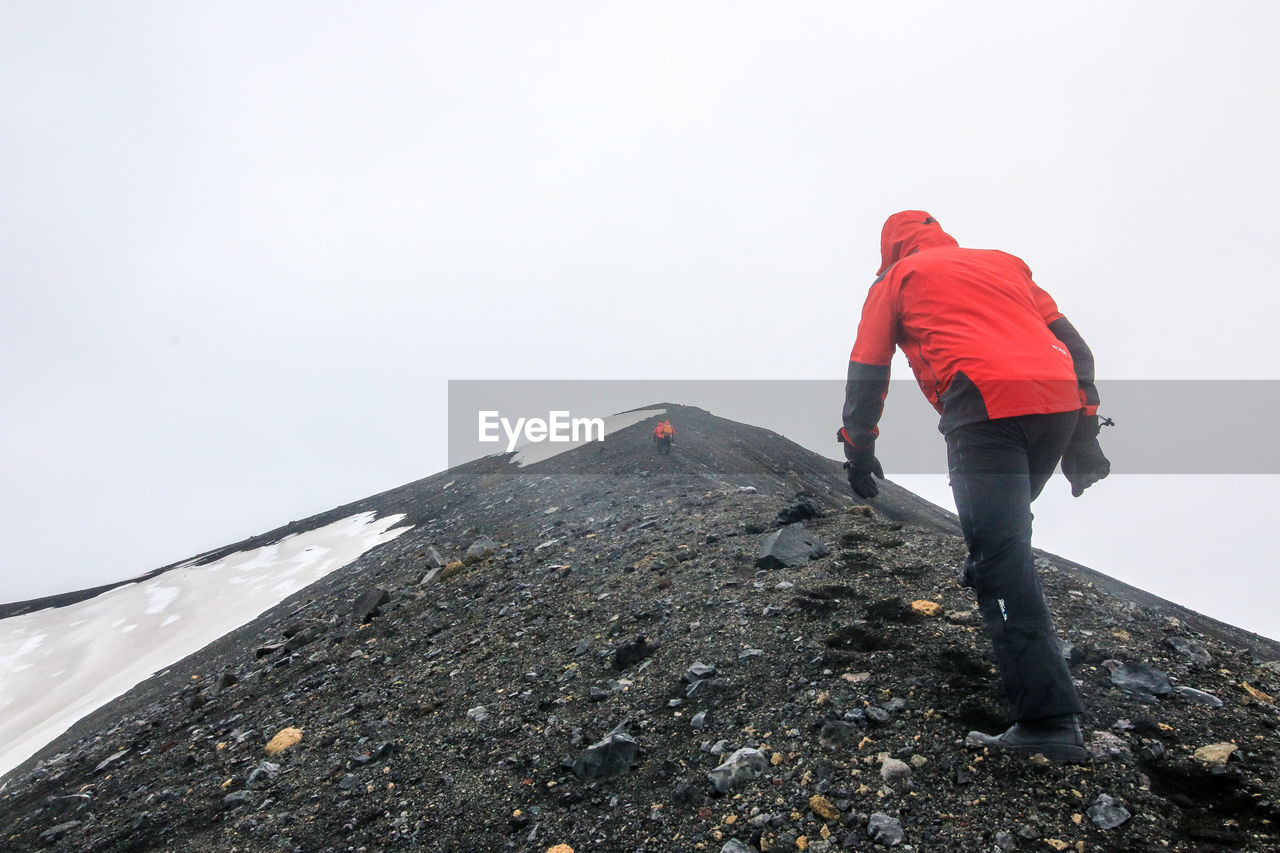 REAR VIEW OF MAN STANDING ON SNOW AGAINST SKY