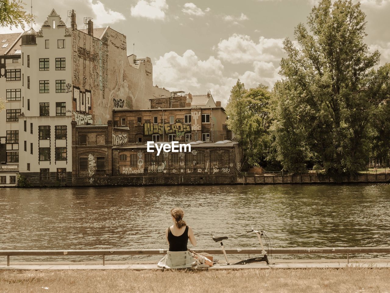 WOMAN SITTING BY RIVER AGAINST BUILT STRUCTURE