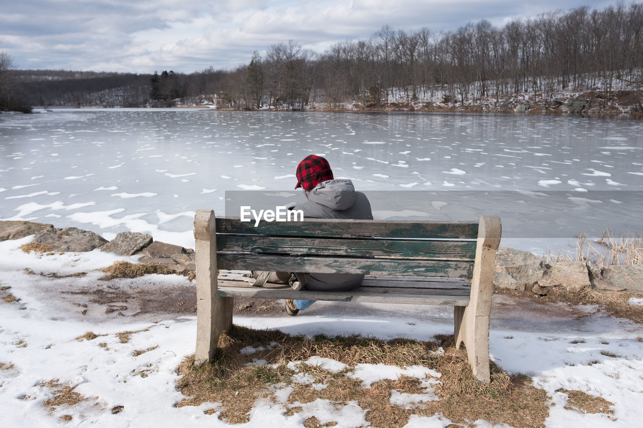 Man sitting on bench by frozen lake against sky during winter