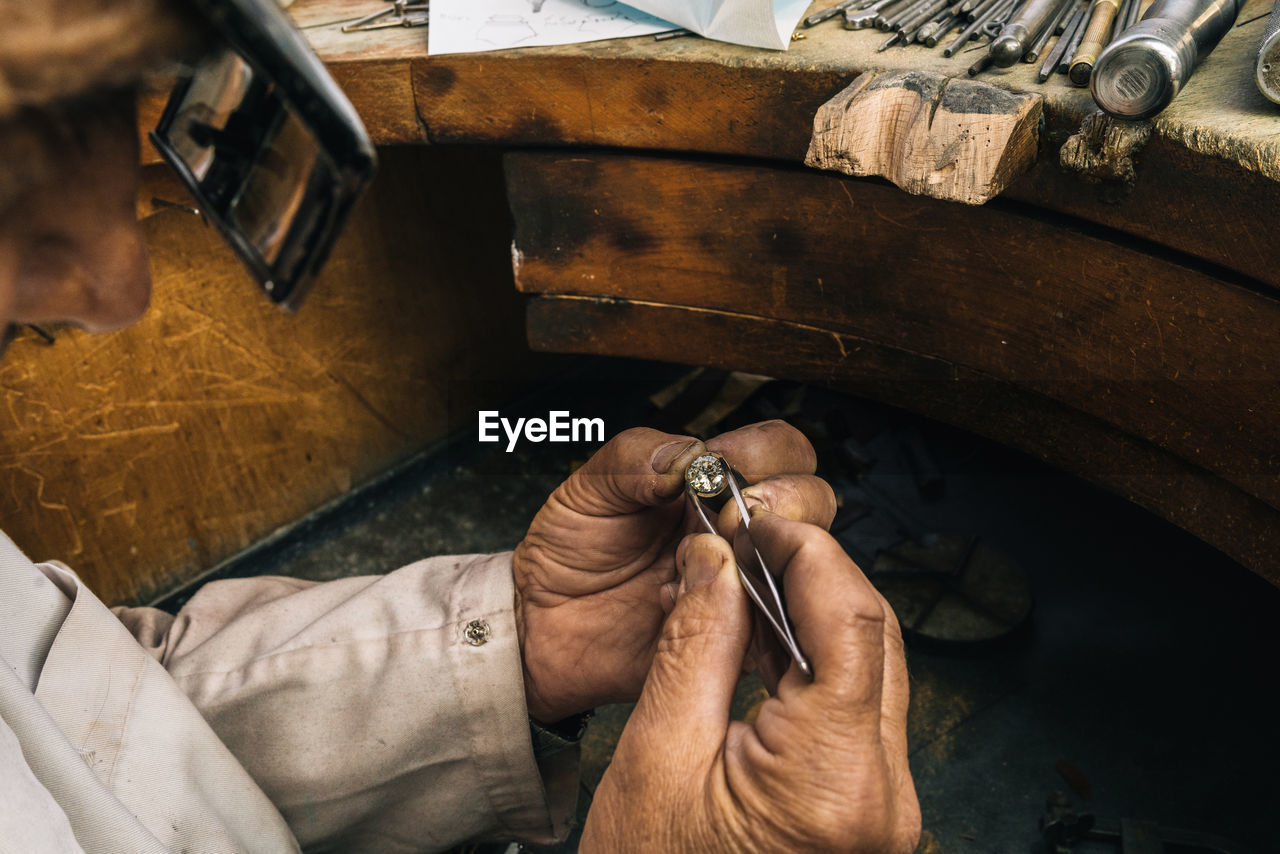 Side view of crop unrecognizable male goldsmith in glasses holding tweezers with diamond while creating jewelry accessory at workbench