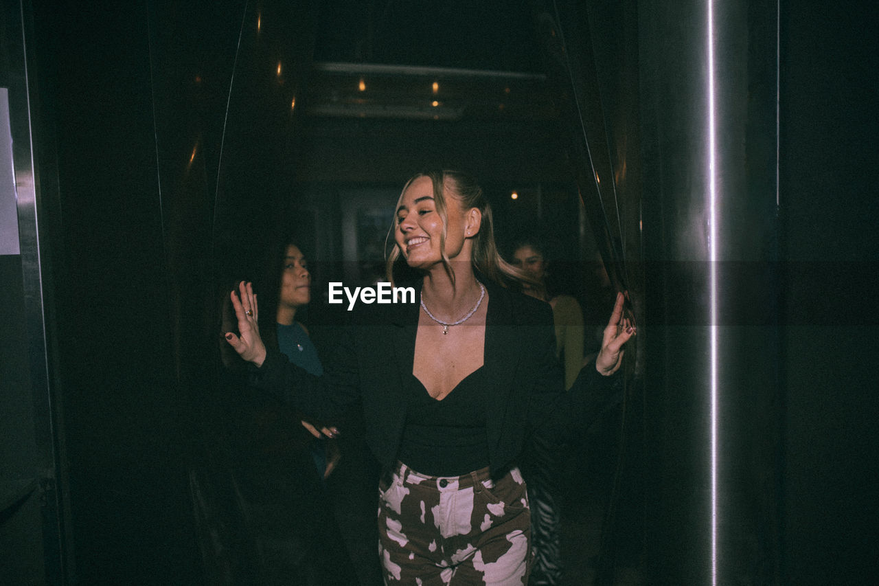 Cheerful young woman getting out of bathroom with friends in background at nightclub