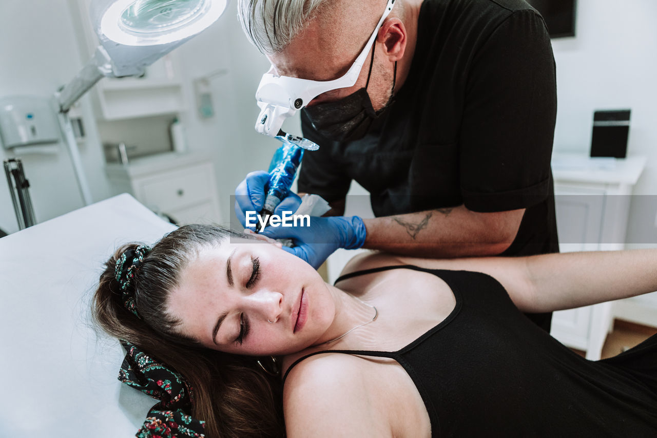 Side view of mature male tattooist in protective mask and medical glasses with tattoo machine making micro tattoo on ear of female customer