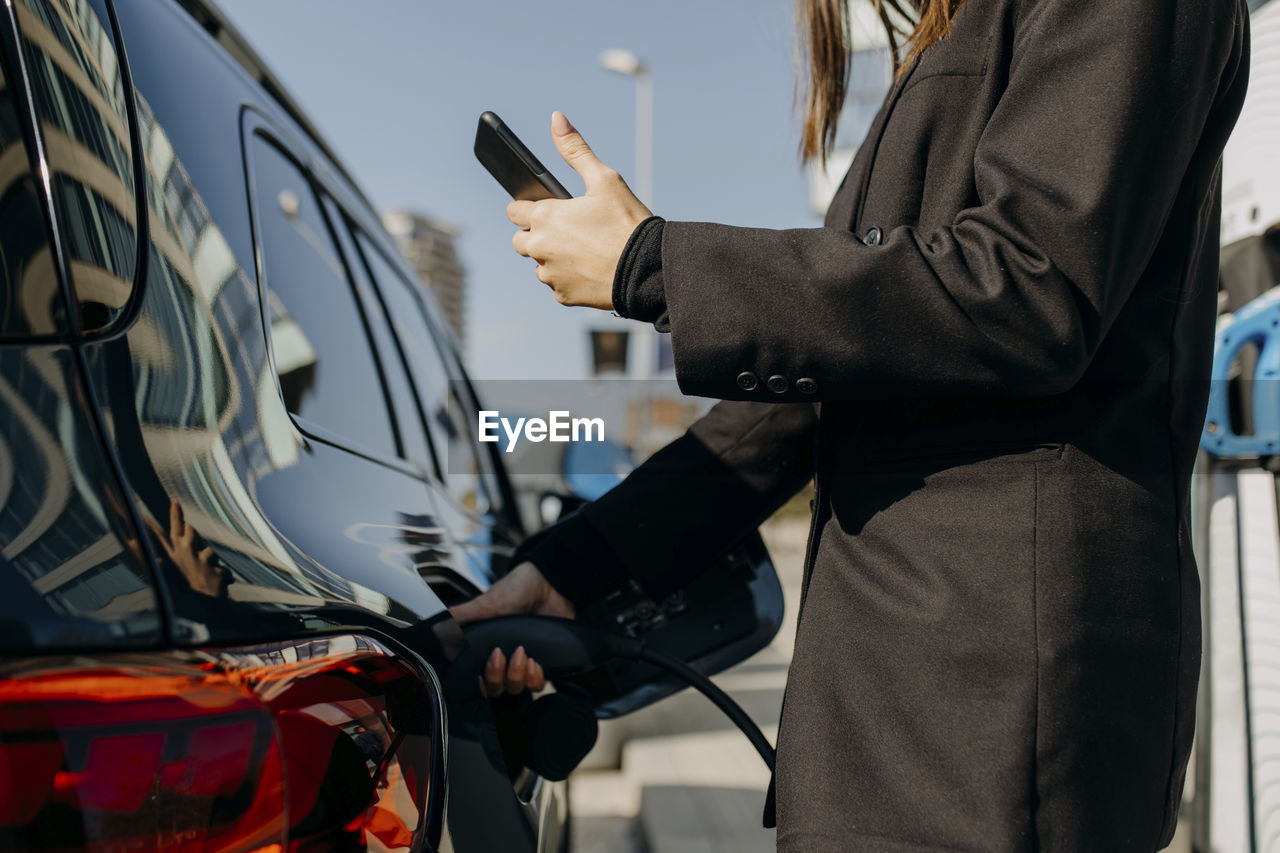 Woman using smart phone near car being charged at charging point