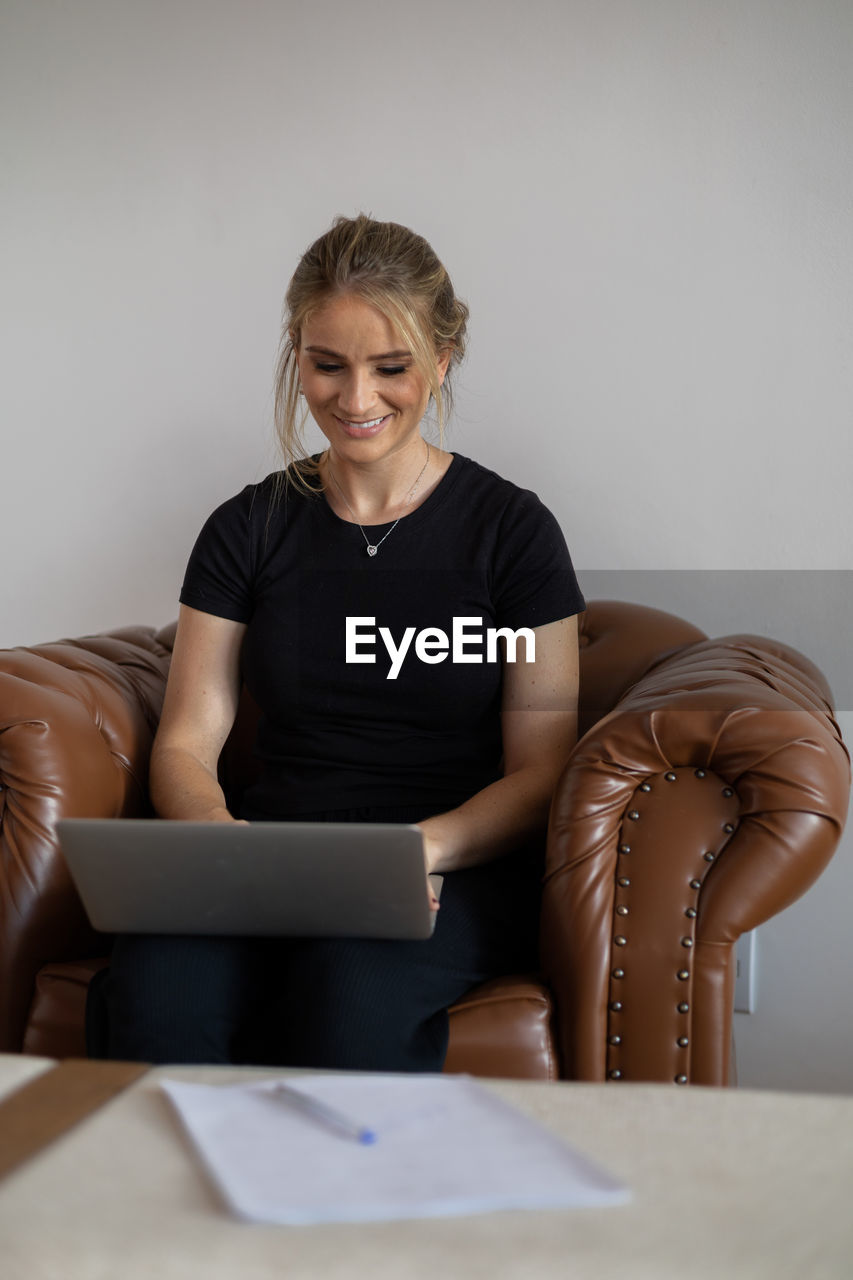 sitting, adult, indoors, women, one person, furniture, front view, clothing, technology, smiling, limb, female, wireless technology, business, person, computer, laptop, communication, portrait, using laptop, businesswoman, sofa, casual clothing, blond hair, mature adult, lifestyles, human leg, happiness, couch, business finance and industry, working, chair, domestic room, emotion, table, looking, conversation, home interior, occupation, hairstyle, three quarter length, copy space, internet, living room