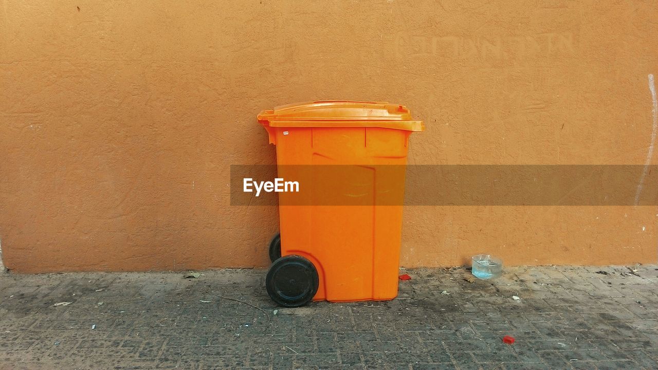 Orange garbage bin on footpath against wall