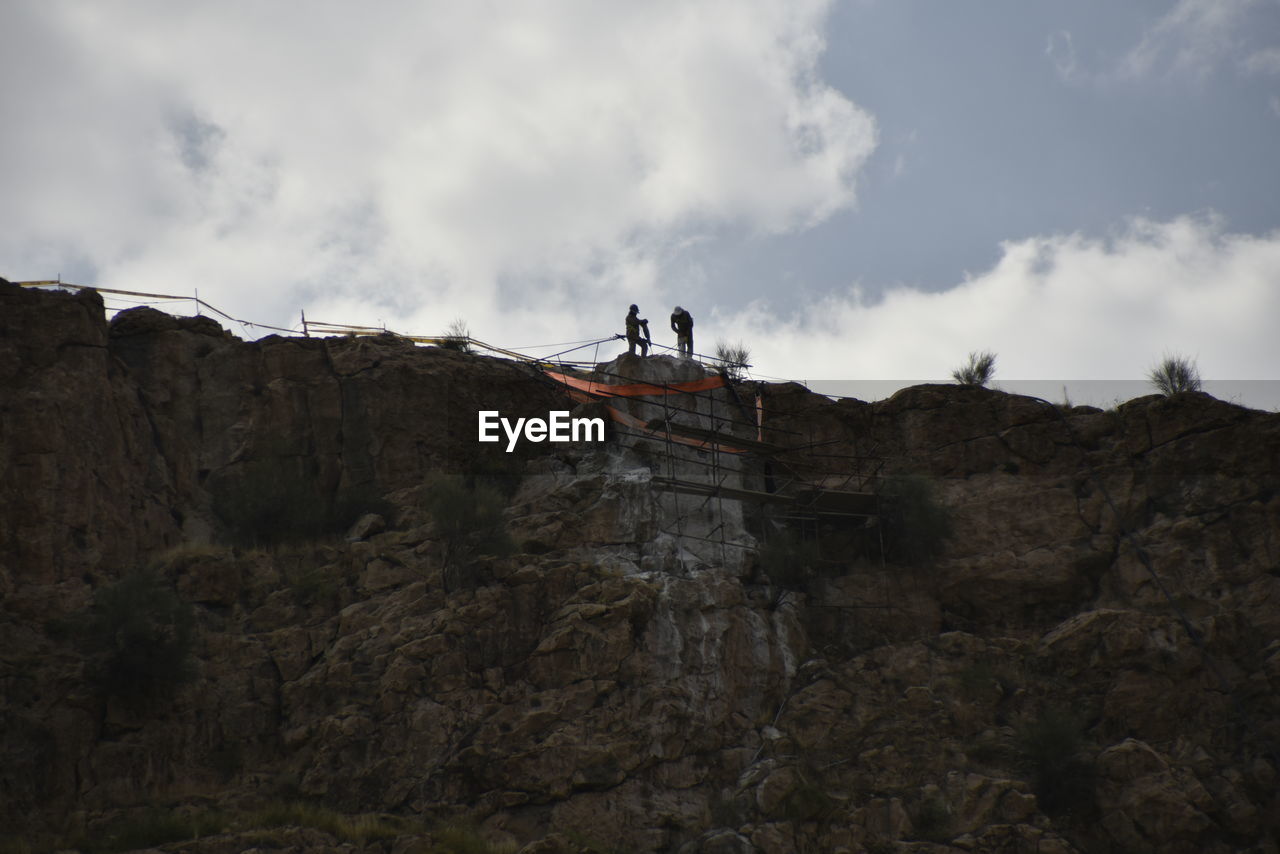 LOW ANGLE VIEW OF PEOPLE ON ROCKS AGAINST MOUNTAIN