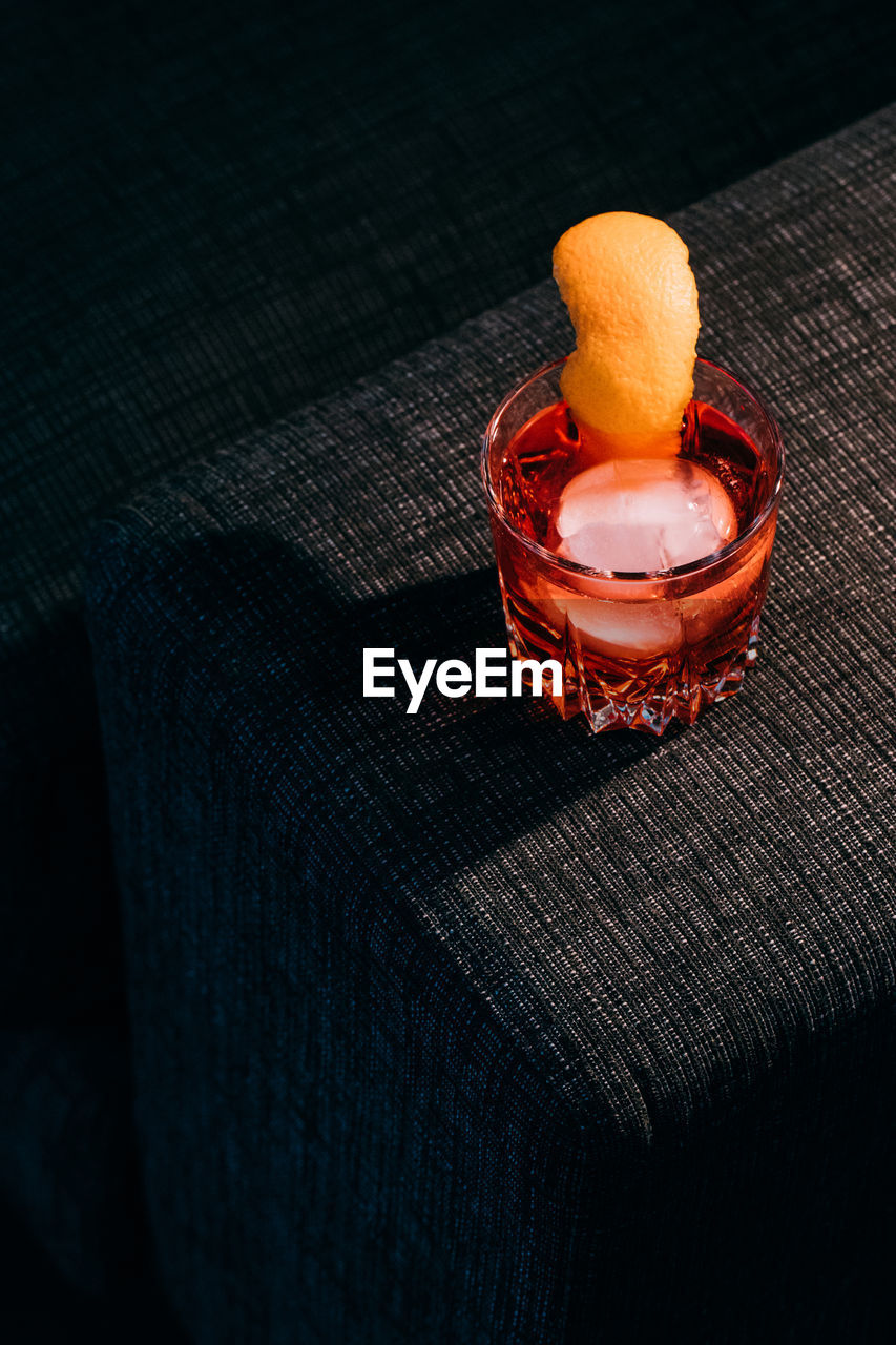 Glass of refreshing negroni cocktail with bitter flavor and ice garnished with orange peel and served on couch arm in dark room