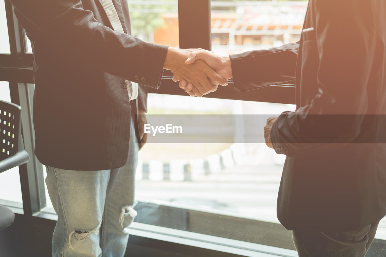 Midsection of businessmen shaking hands while standing by window in office