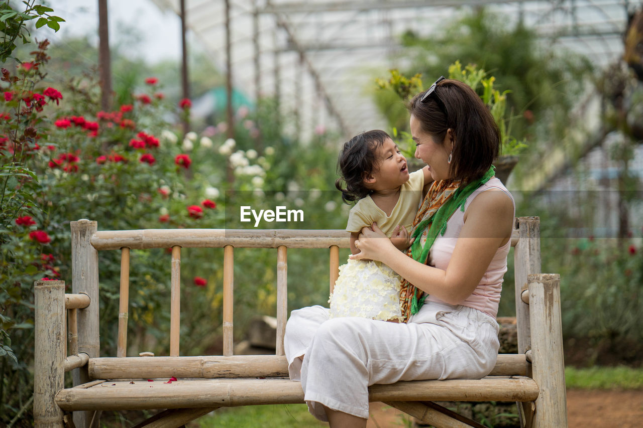 Side view of mother and daughter sitting on bench by plants